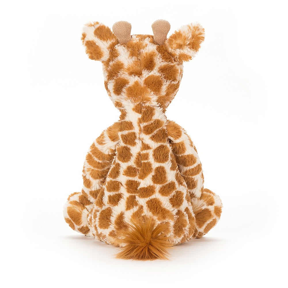 Soft plush Bashful Giraffe toy with a spotted brown and cream pattern, sitting down facing away, showcasing its fluffy tail and gentle features.