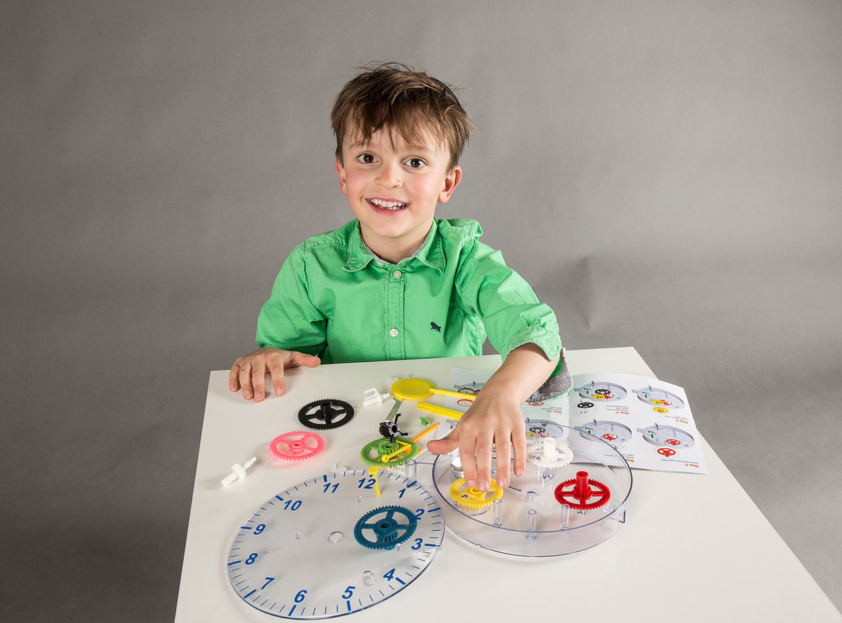 A young boy in a green shirt smiles while sitting at a white table with a DIY Clock kit. The kit includes a clear round base, colorful dials in blue, red, yellow, green, and black, and an instruction sheet on a gray background.