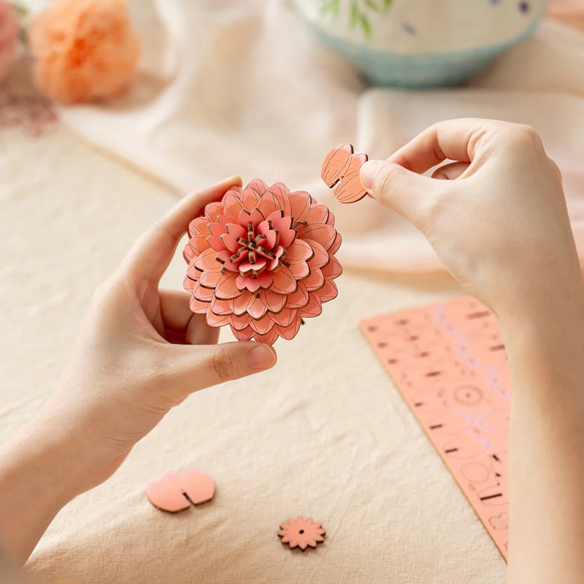 Person assembling a Pink Dahlia DIY Wooden Flower 3D Puzzle with craft materials on a table.