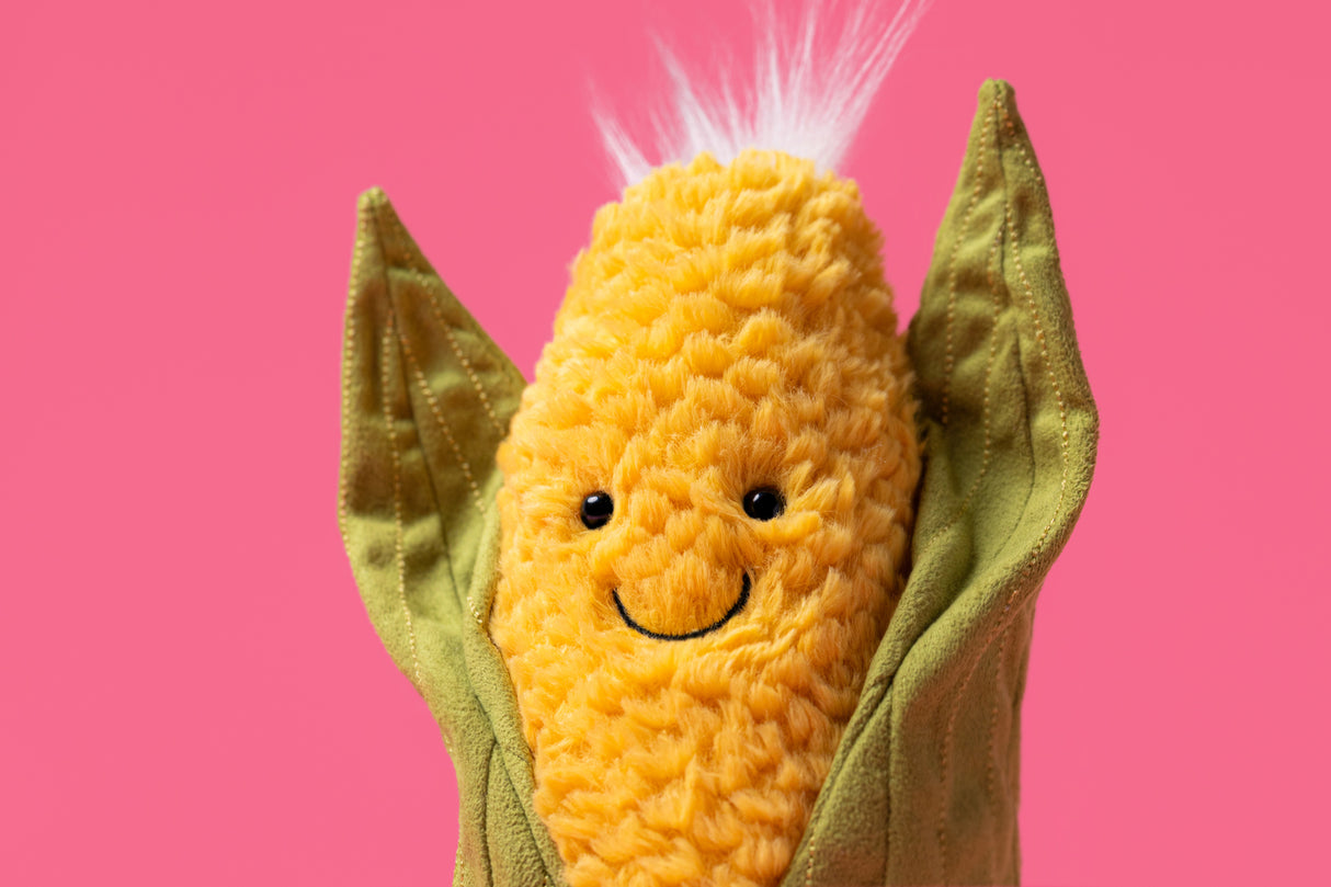Amuseables Sweetcorn plush toy with a smiling face, soft yellow textured body, green fabric husk, and fluffy white top against a pink background.