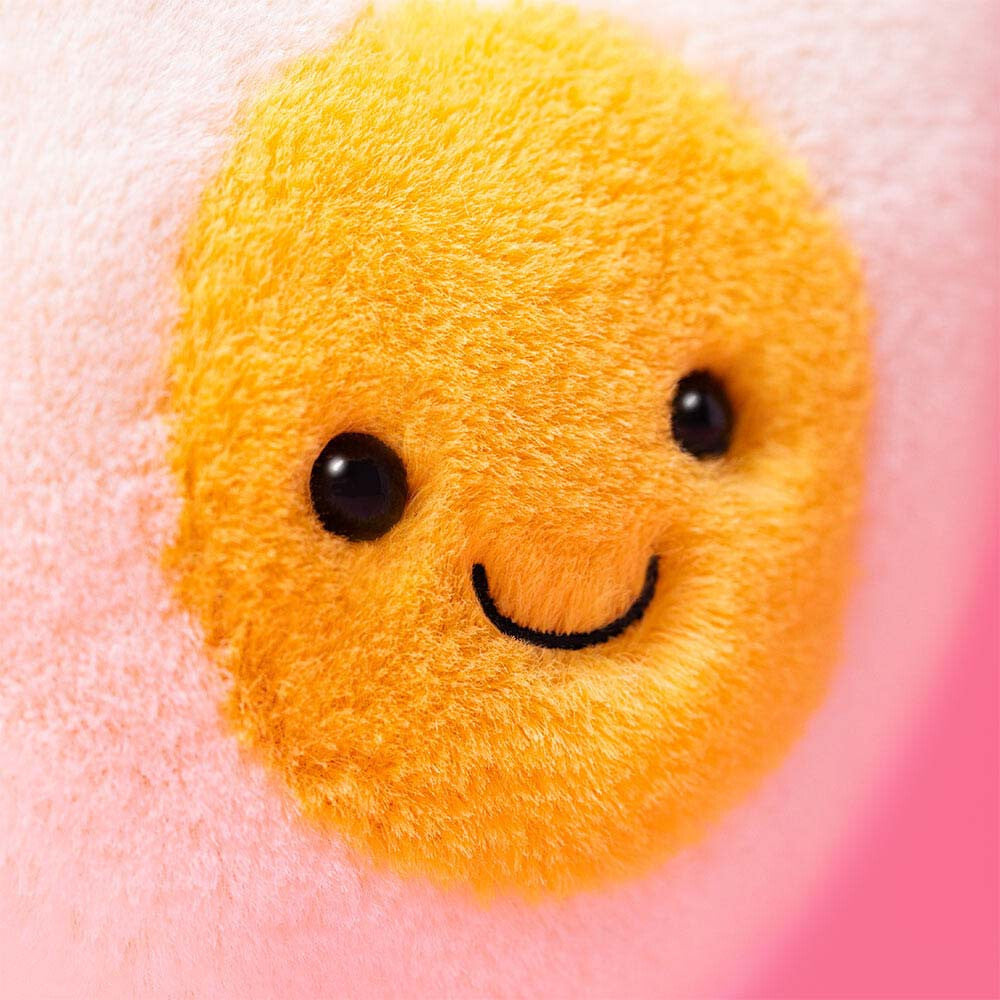 Close-up of the smiling face on the soft yellow center of the Amuseables Heart Boiled Egg plush toy with a pink background.
