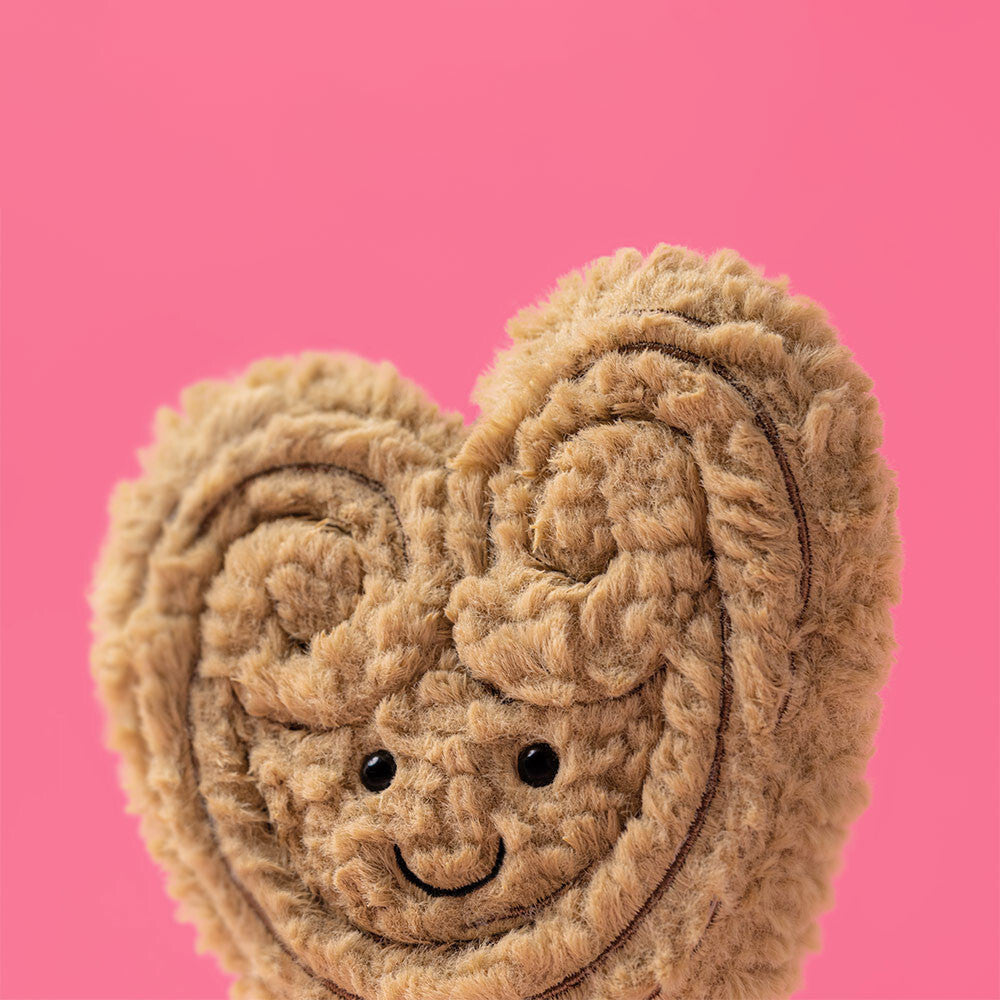 Soft plush toy shaped like a heart with a smiling face and textured details in light brown, named Amuseables Philippe Palmier, against a pink background.