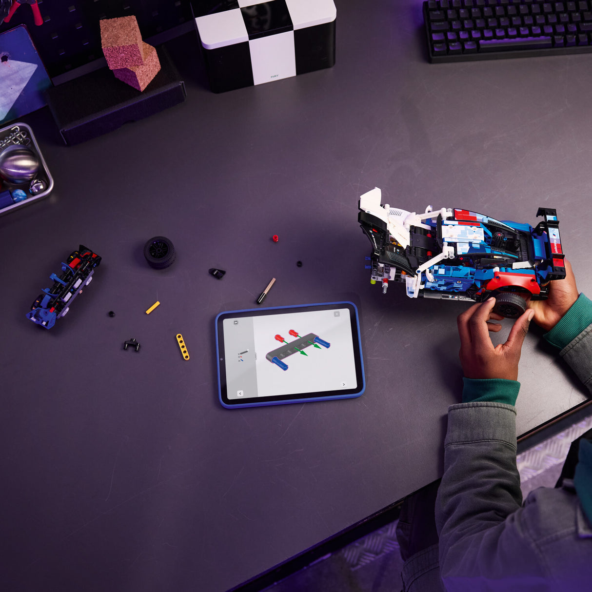 Person assembling a LEGO Technic: BMW M4 GT3 EVO Race Car model on a desk with a tablet displaying design software.