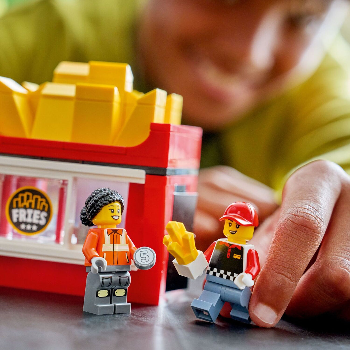 Child playing with LEGO City Great Vehicles: Fries Food Truck figures in front of a toy fast food restaurant set.