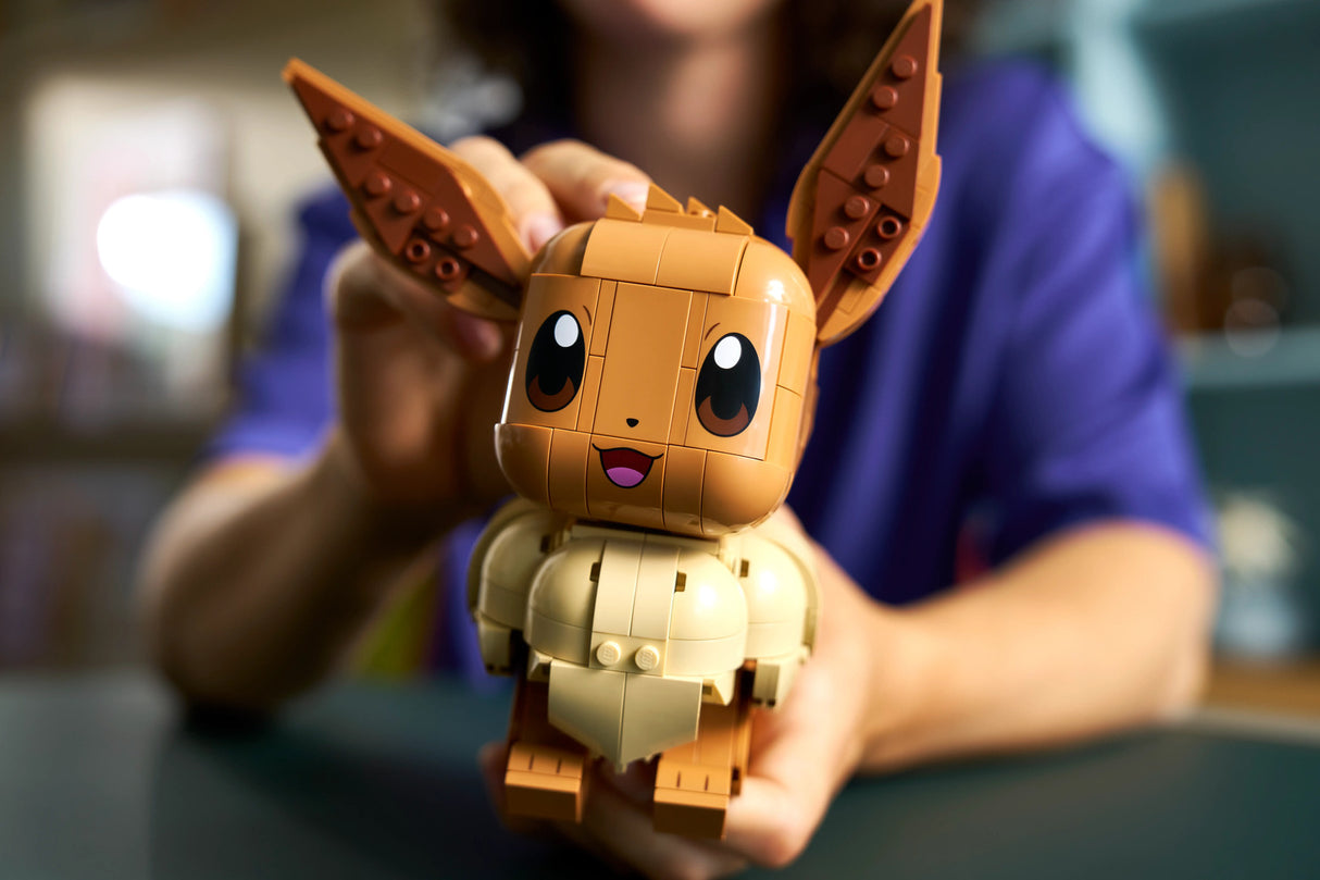 A toy building block figure of a cute LEGO Pokémon Eevee with large pointed ears, held up by a person wearing a blue shirt in a softly blurred indoor setting.