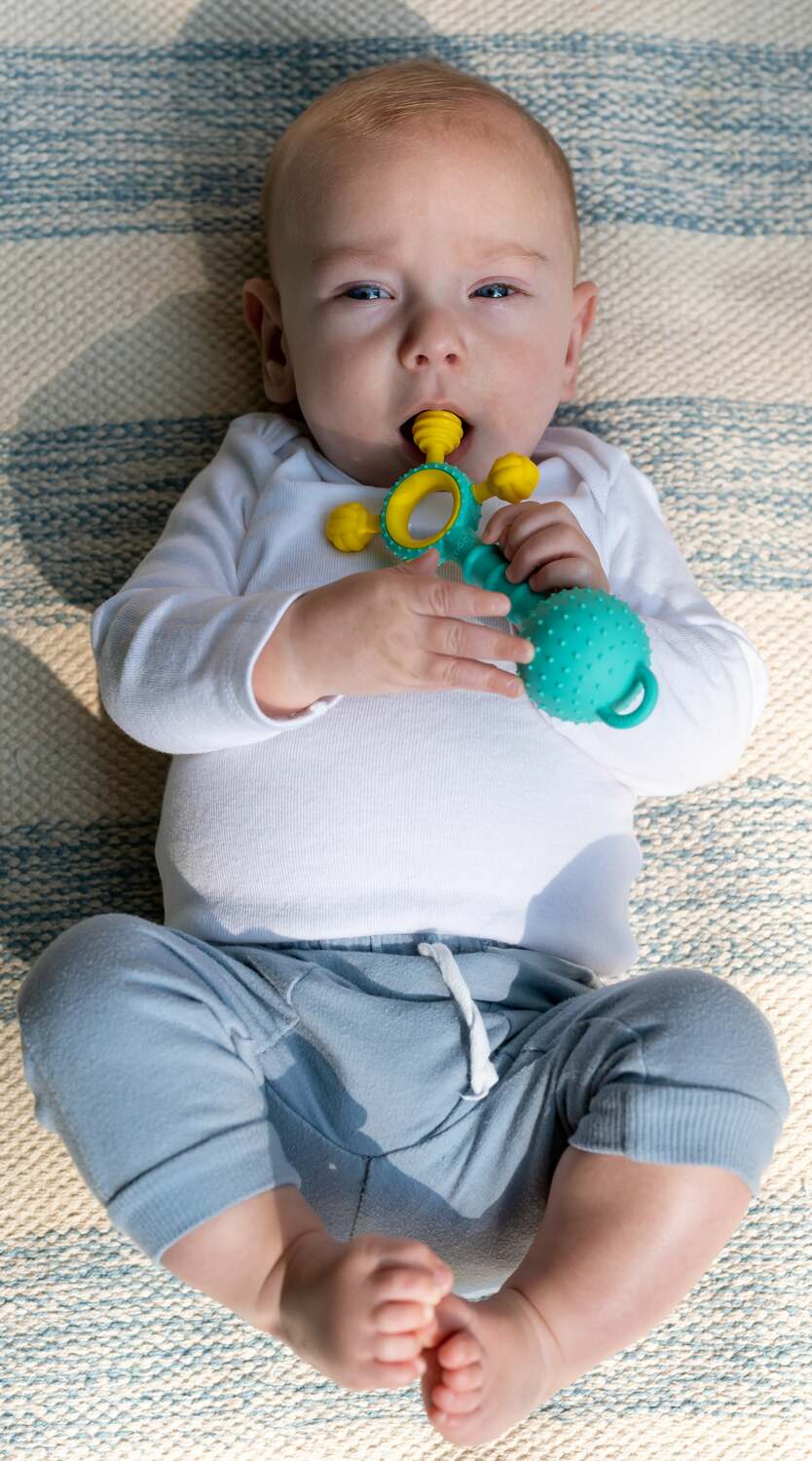 Baby lying on a striped blanket holding a Gumlii Teether Rattle
