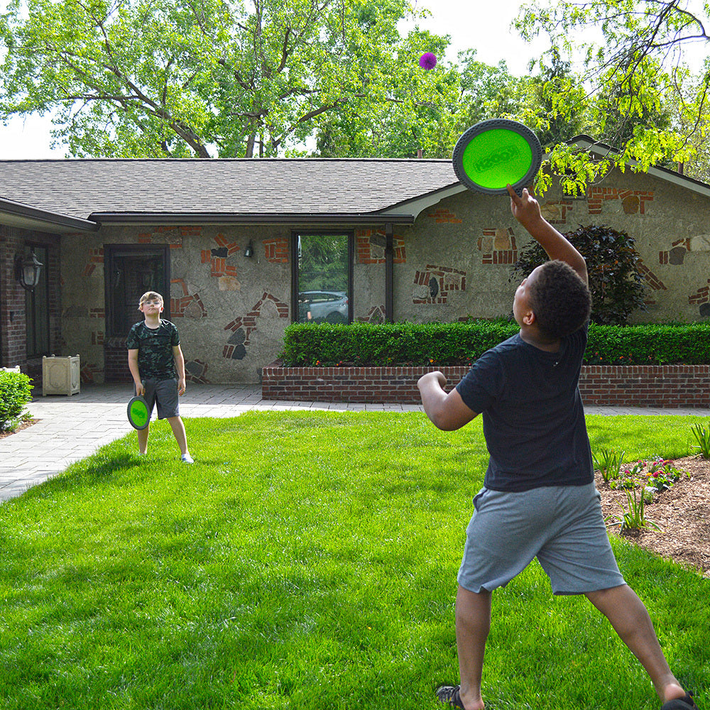 Two children playing with Koosh® Paddle Play Set in a backyard with a house in the background.