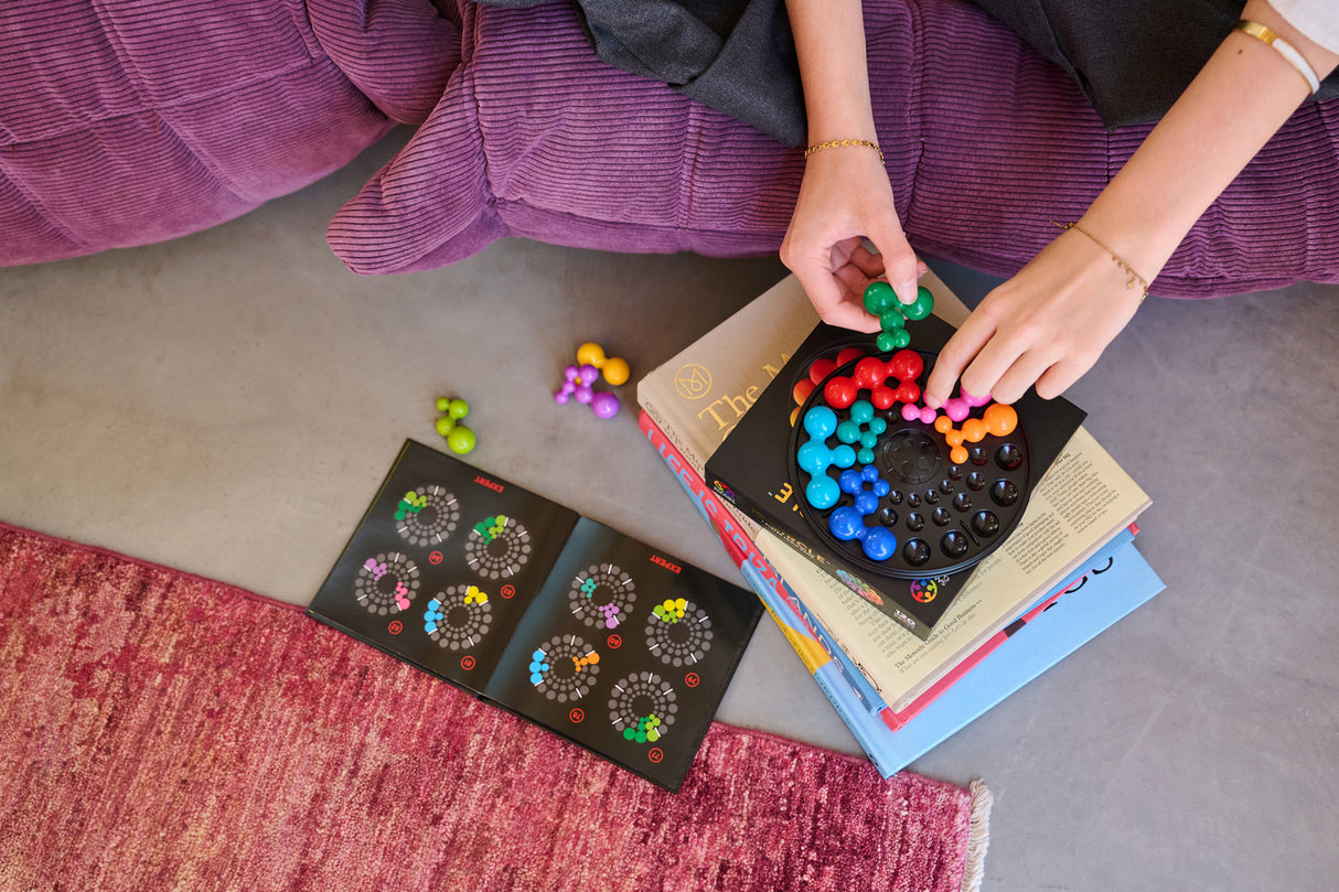 Person playing with a colorful IQ Deluxe - Circle puzzle on a stack of books, sitting on purple pillows.