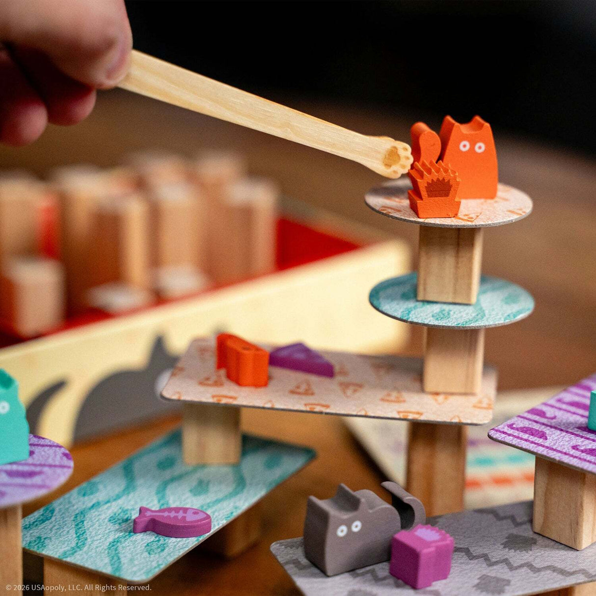 A close-up of a wooden Cats Knocking Things Off Ledges Balance Game being built with a hand holding a wooden stick. The toy features colorful animal-shaped pieces and patterned platforms on natural wood posts.