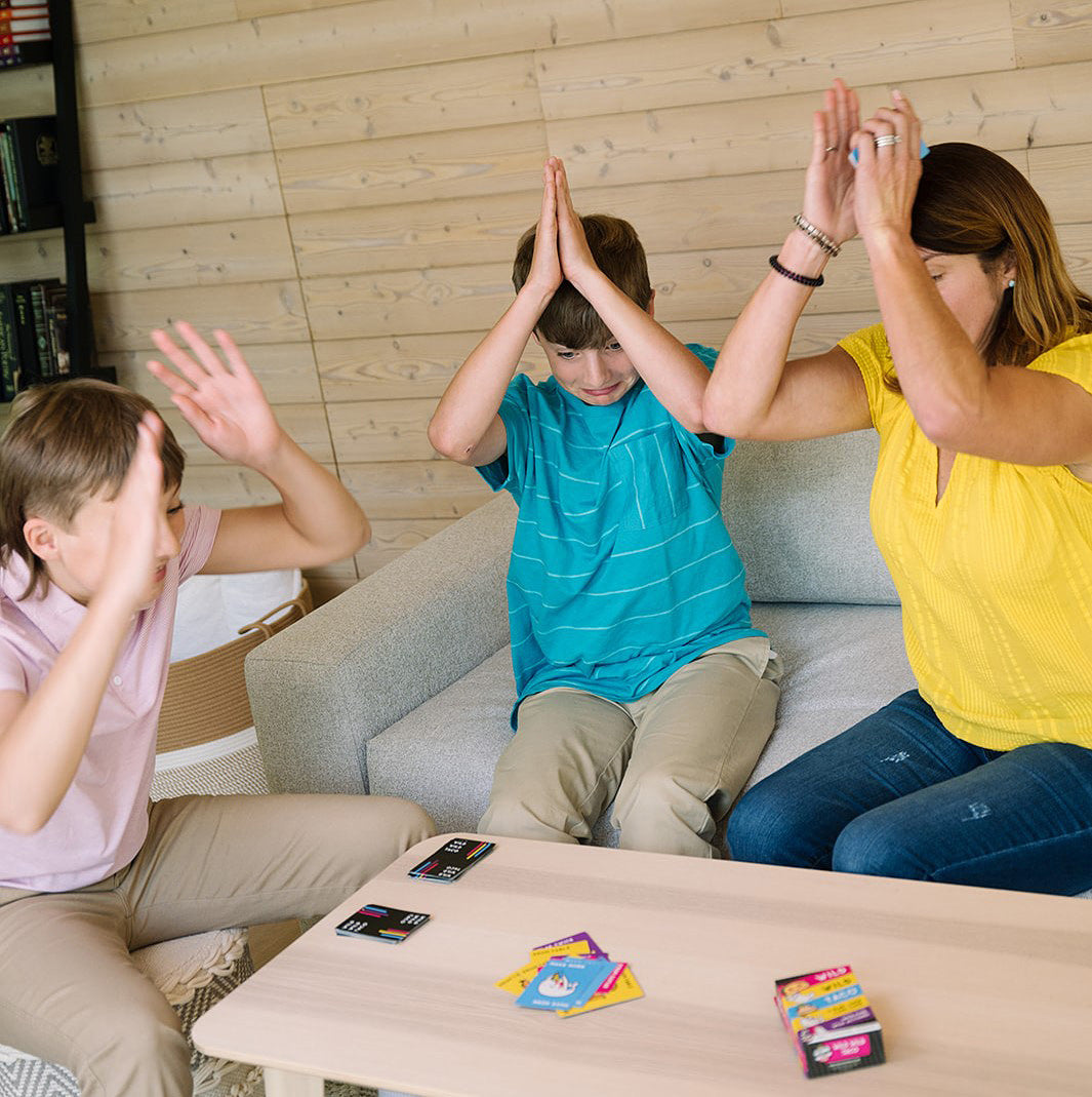 Three people sitting on a couch with their hands raised, playing Wild Wild Taco Card Game on a coffee table.