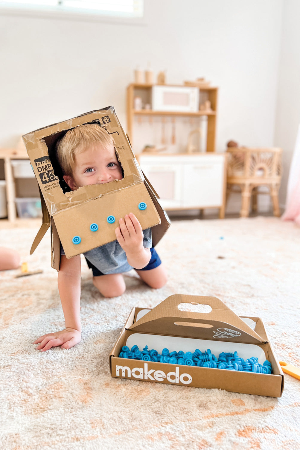 Child playing with cardboard pieces and a box labeled 'makedo' on a carpeted floor.