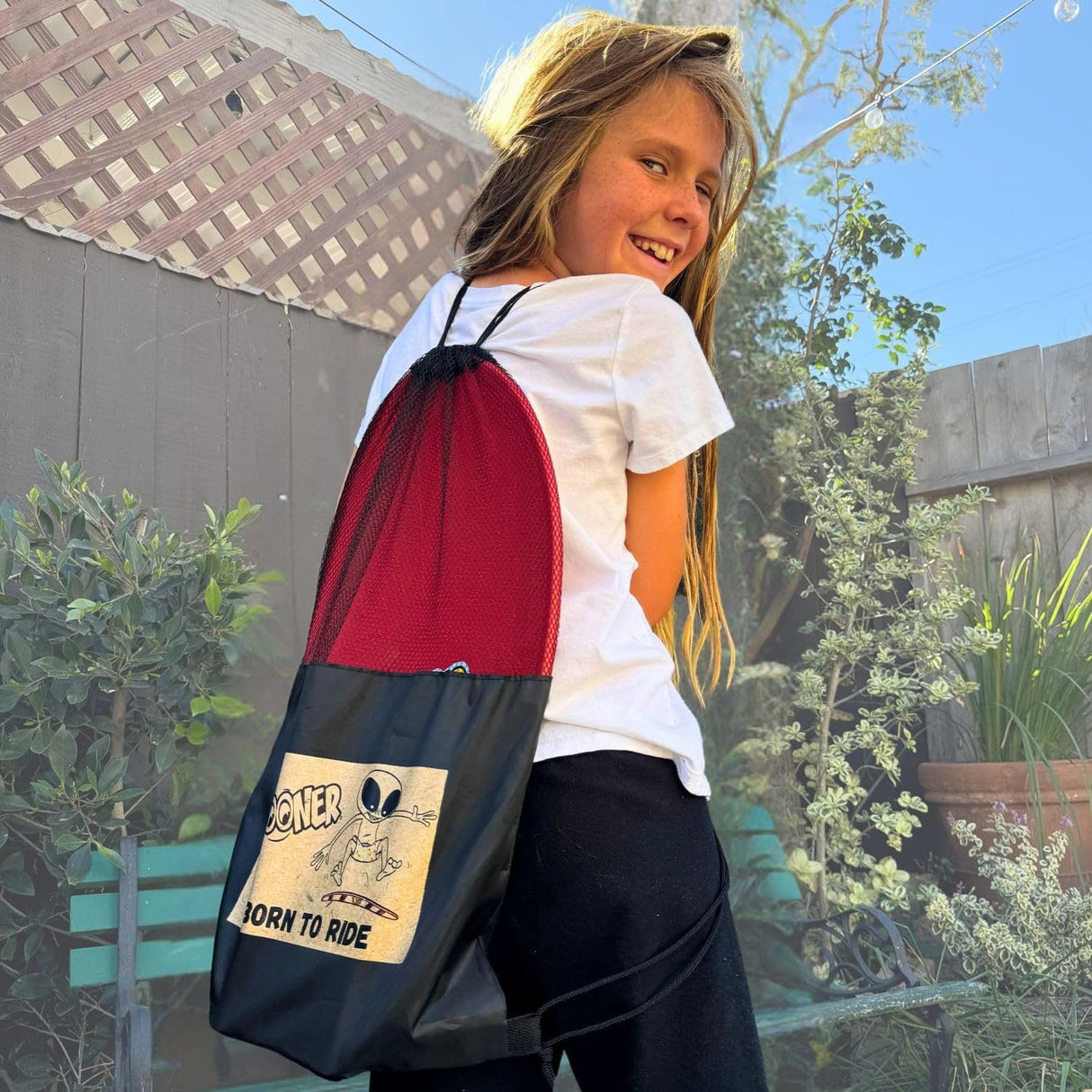 Child carrying a The Spooner Pro Born to Ride Training System - 26" in a red and black drawstring bag with a graphic design outdoors.