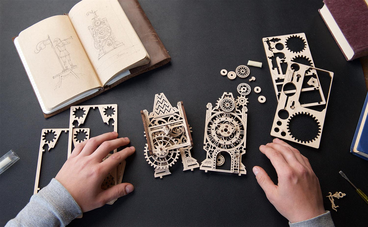 A person's hands are assembling a  wooden Old Clock Tower model with gears and cogs on a dark tabletop, surrounded by laser-cut wooden panels, a sketchbook, and other parts.