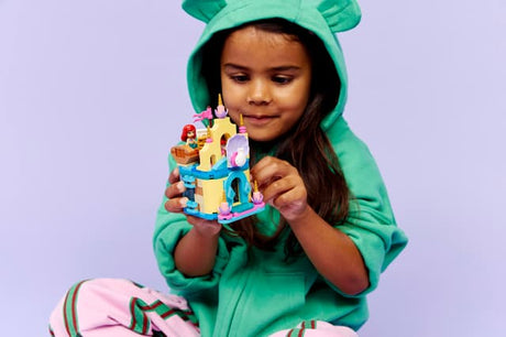 Child in a green hoodie holding a LEGO Disney Princess: Ariel's Magical Mini Palace against a light purple background