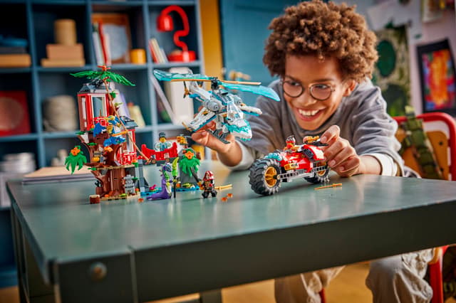 Child playing with LEGO Ninjago: Ninja Vehicle Tree House Battle set on a table in a room with shelves and toys.