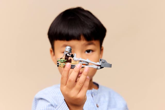 Child holding a LEGO Star Wars: The Mandalorian & Grogu's Speeder Bike™ in front of their face against a beige background