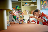 Two children playing with a LEGO Super Heroes Marvel: Spider-Man vs. Mysterio: The Daily Bugle set at a table in a room with shelves in the background.