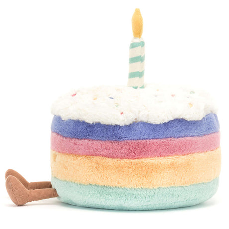Soft plush toy designed as a colorful birthday cake with rainbow layers, white frosting sprinkled with confetti, a striped candle on top, and small legs, named Amuseables Rainbow Birthday Cake.