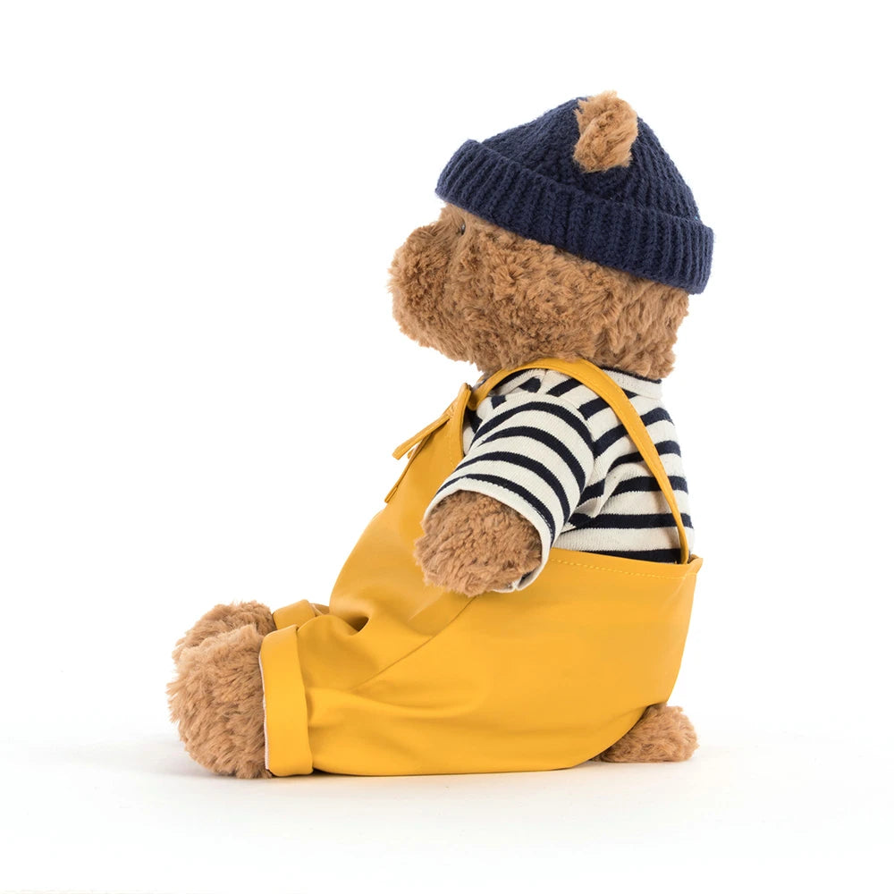 Soft brown teddy bear dressed in a striped shirt, navy knit hat, and bright yellow overalls wearing the Bartholomew Bear Seafarer Outfit, perfect for kids dress-up and plush collections.