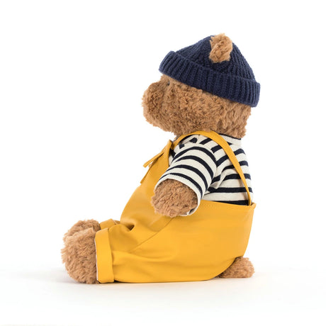 Soft brown teddy bear dressed in a striped shirt, navy knit hat, and bright yellow overalls wearing the Bartholomew Bear Seafarer Outfit, perfect for kids dress-up and plush collections.