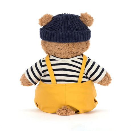 Bartholomew Bear Seafarer Outfit dressed teddy bear wearing a navy blue knit hat, striped shirt, and mustard yellow overalls , viewed from the back.