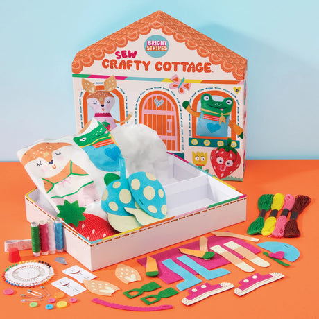 Bright Stripes Sew Crafty Cottage kit displayed open with its house-shaped box, fabric pieces, embroidery floss, beads, and other sewing supplies arranged on an orange surface against a light blue background.