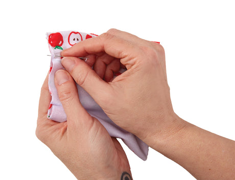 Close-up of a person's hands pinning a white fabric piece with a red and white apple pattern, set against a solid white background.