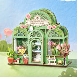 Green Fountain Garden, Super Mini Houses with glass panels, pink roses, small potted plants, a white fountain centerpiece, and decorative garden accessories, shown against a soft blue sky and green grass background.