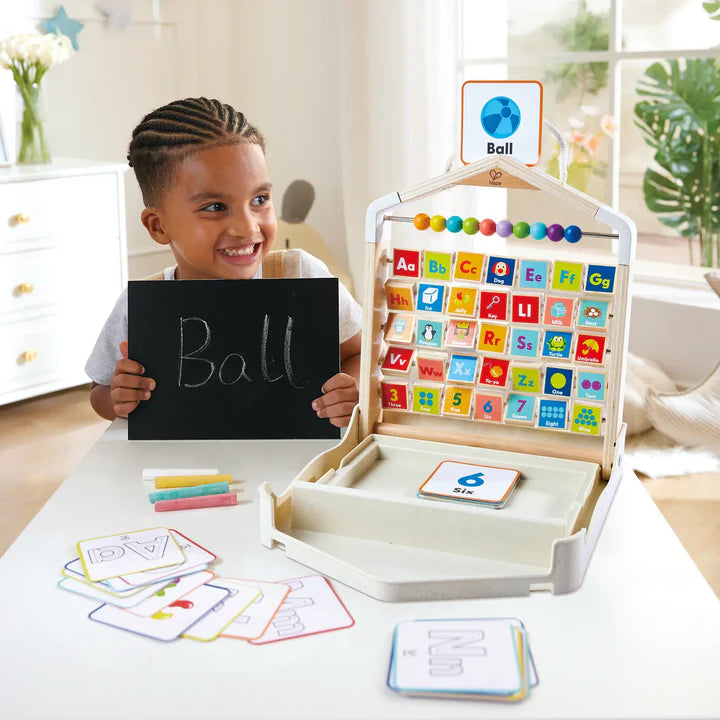 Child holding a blackboard with 'Ball' written on it, next to an educational Alphabet Learning Pack with letters and numbers.
