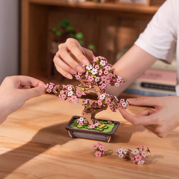 Sakura Bonsai DIY Wooden 3D Puzzle with pink flowers being held by hands on a wooden surface.