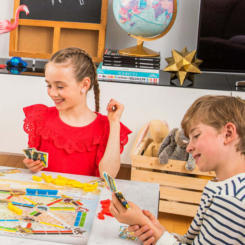 Two children playing Ticket to Ride: First Journey at a table with educational items in the background.