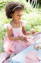 Young girl in a pink dress sitting on grass wearing Boutique Heartbreaker Headband, surrounded by nature.