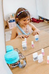 Young girl wearing a Boutique Heartbreaker Headband  and playing with miniature nail polish bottles on a wooden surface.