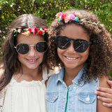 Two young girls wearing sunglasses and flower crowns outdoors.