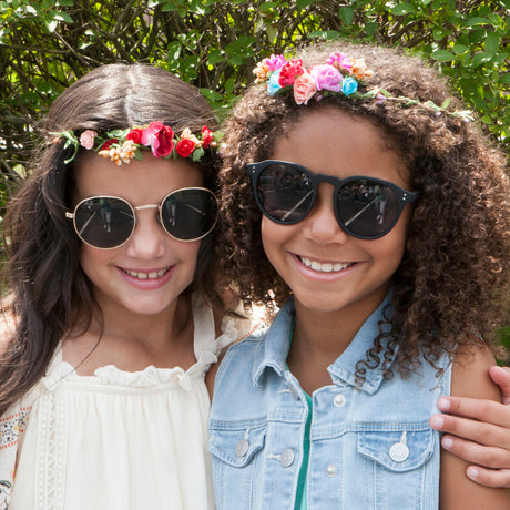 Two young girls wearing sunglasses and flower crowns outdoors.