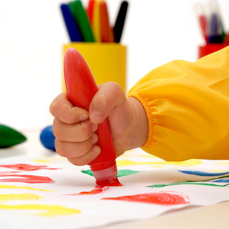Child's hand holding a red Little Creatives Jumbo Washable Squeeze Paintbrushe with colorful crayons and a yellow container in the background.