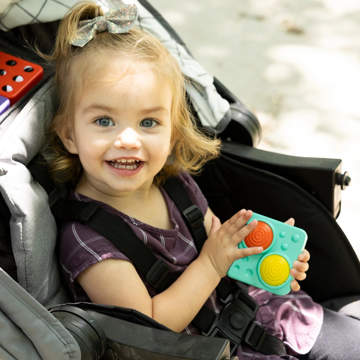 Child in a stroller holding a colorful PlayTab Tiles Set 4 , smiling outdoors.