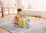 Child playing with a colorful Green Thumbs Activity Cube on a rug in a room with shelves and a couch.