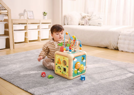 Child playing with a colorful Green Thumbs Activity Cube on a rug in a room with shelves and a couch.