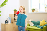 A young girl stands in a bright, modern room holding a Popstar Microphone (Pixelz). She is wearing a sleeveless teal knit cardigan over a cream long-sleeve shirt and blue jeans.