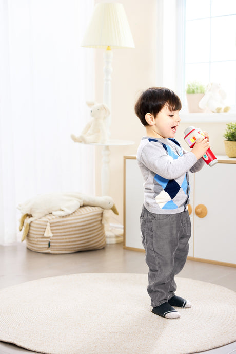 A young boy stands on a round cream rug in a bright, modern nursery, holding a red and white Popstar Microphone (Pixelz). He is wearing a gray long-sleeve shirt, a blue and gray argyle sweater vest, and gray pants.
