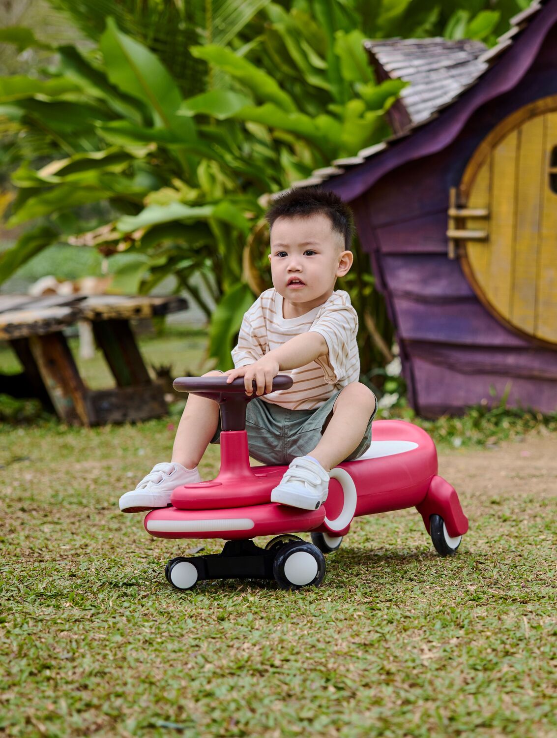 Child sitting on a Wiggle & Giggle Car, Red in an outdoor setting with greenery and a small playhouse.