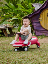 Child sitting on a Wiggle & Giggle Car, Red in an outdoor setting with greenery and a small playhouse.