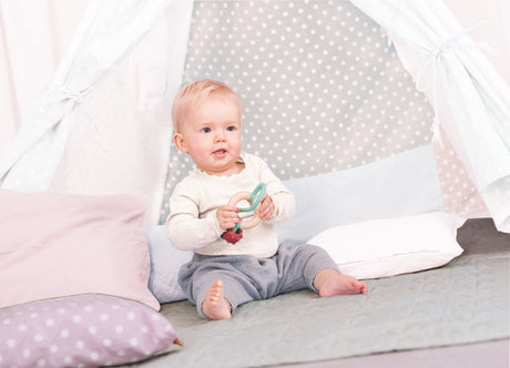 Baby sitting on a bed and holding a Bead Maze with a crib in the background