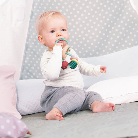 Baby sitting on a bed with a Bead Maze teething ring and toy, against a polka dot curtain background.