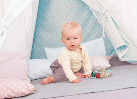 Baby sitting on a bed with colorful Bead Maze , surrounded by soft bedding and pillows.