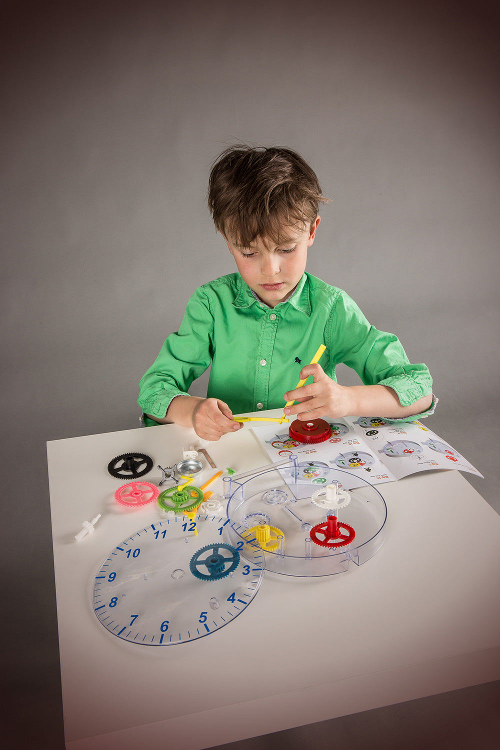 A young boy in a green shirt sits at a table, working on a colorful DIY Clock set with gears, a clear circular tray, and an instruction booklet against a dark gray studio background.