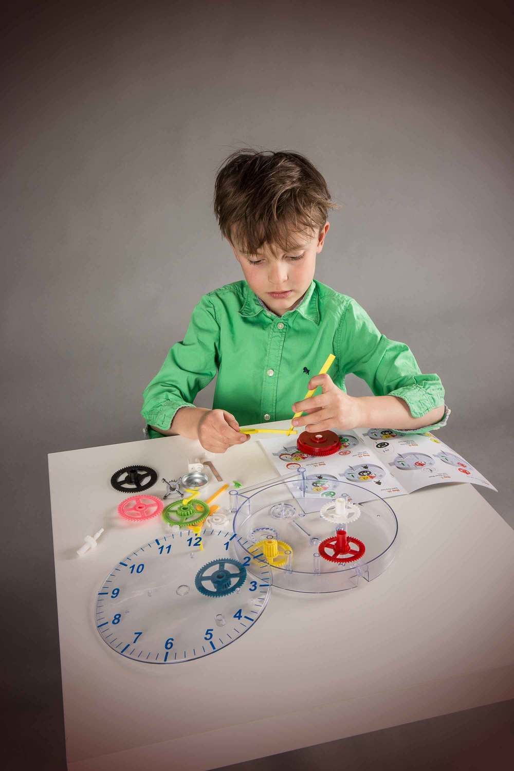 A young boy in a green shirt sits at a table, working on a colorful DIY Clock set with gears, a clear circular tray, and an instruction booklet against a dark gray studio background.