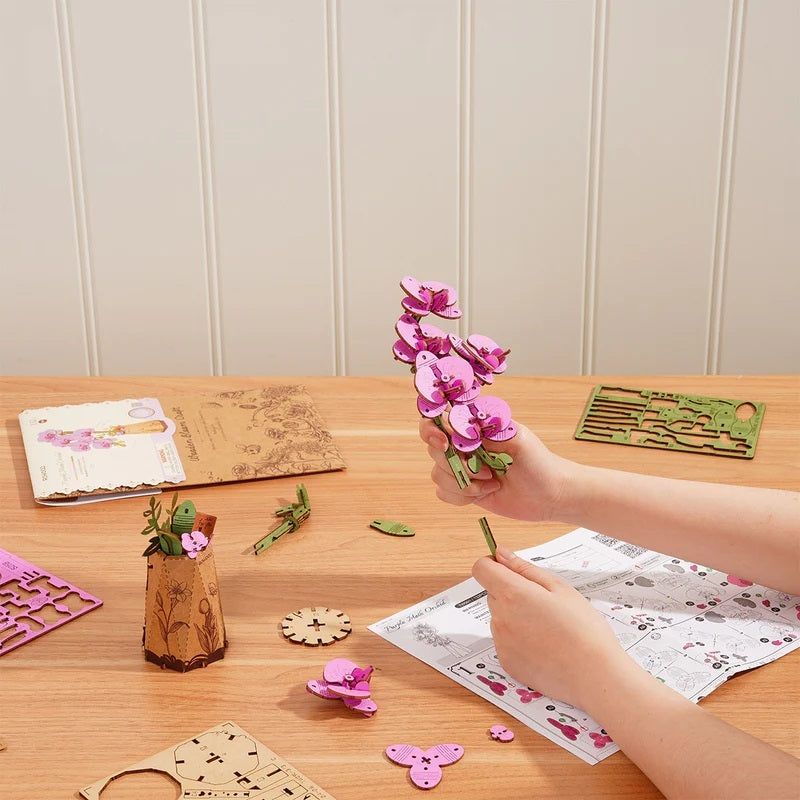 Person assembling Purple Moth Orchid DIY Wooden Flower on a wooden table with craft materials.