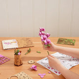 Person assembling Purple Moth Orchid DIY Wooden Flower on a wooden table with craft materials.